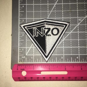 May include: A triangular embroidered patch featuring the text INZO in black and white. The patch is split vertically into a black left side and a white right side, with a thick white border. A pink ruler shows the patch is approximately 3.5 inches wide.