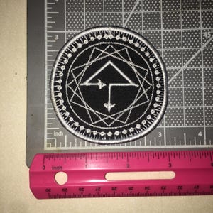 May include: A circular black embroidered patch featuring a white geometric design with a central arrow-like symbol enclosed by a star-shaped border. The patch measures approximately 2.5 inches in diameter as shown by the visible measuring tool.