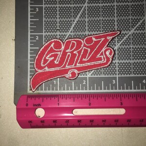 May include: A red embroidered patch with white stitching featuring the text GRIZ in a stylized font. The patch is positioned on a cutting mat next to a pink ruler, showing the design measures approximately 3.5 inches in total width.