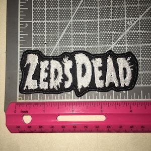 May include: A black embroidered patch with white text reading ZEDS DEAD in a distressed, jagged font. The patch is positioned above a pink plastic ruler, showing the item measures approximately 4.25 inches in total length from end to end.