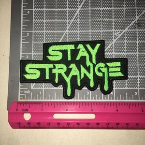 May include: A black embroidered patch with the text STAY STRANGE in bright green, featuring a dripping font style. The patch is positioned on a cutting mat next to a pink ruler, showing a width of approximately 4.5 inches and a height of 2.5 inches.