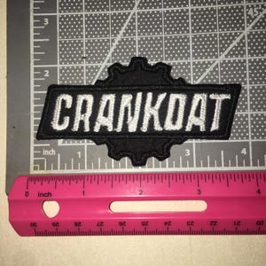 May include: A black embroidered patch featuring the white text CRANKDAT centered over a gear-shaped background. The patch is positioned on a cutting mat with a pink ruler, showing the patch is approximately 3.5 inches wide and 1.5 inches tall.