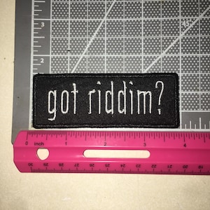 May include: A rectangular black embroidered patch featuring the white text "got riddim?" in a lowercase sans-serif font. The patch is positioned above a pink ruler, showing a width of approximately 4 inches and a height of 1 inch.