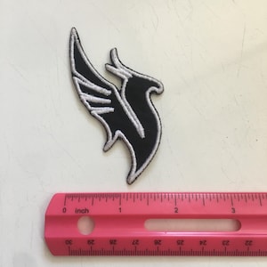 May include: An embroidered patch featuring a stylized black bird silhouette with white stitched outlines. The patch is positioned above a pink plastic ruler that shows the item measures approximately 3.5 inches in total height and width.