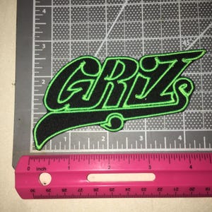 May include: A black embroidered patch with bright green stitching outlining the text GRIZ. The patch is positioned on a cutting mat next to a pink ruler, showing the design measures approximately 4.5 inches in total width and 2 inches in height.