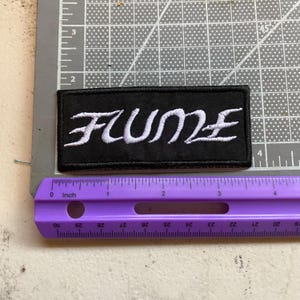 May include: A rectangular black embroidered patch featuring the white text Flume in a stylized font. The patch is positioned above a purple ruler, showing a width of approximately 4 inches and a height of approximately 1.5 inches.
