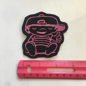 May include: A black embroidered patch featuring a cartoon character wearing a baseball cap and a striped shirt, shown next to a pink ruler that indicates the patch is approximately 3 inches wide and 3 inches tall in total dimensions.