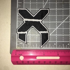 May include: A black embroidered patch in the shape of the letter X with white stitched borders and two horizontal white stripes across the center. The patch is positioned on a cutting mat next to a pink ruler showing a width of 2.5 inches.
