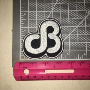 May include: A black and white embroidered patch featuring a stylized, interconnected letter design. The patch is positioned on a cutting mat next to a pink ruler, showing the design measures approximately 2.5 inches in width and height.