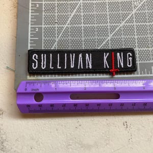 May include: A rectangular black embroidered patch featuring the white text SULLIVAN KING with a red sword icon replacing the letter I. The patch is positioned above a purple ruler, showing a length of approximately 4 inches on the imperial scale.