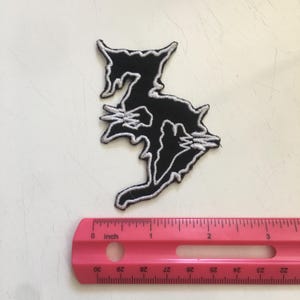 May include: A black embroidered patch with a jagged, stylized shape outlined in white thread. The patch is positioned above a pink plastic ruler that shows the object is approximately 3.5 inches tall and 2.5 inches wide at its widest points.