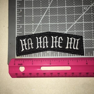 May include: A black embroidered patch with white text reading HA HA HE HU in a gothic font. The patch is positioned above a pink plastic ruler, showing the item is approximately 4.5 inches wide and 1.25 inches tall at its highest point.