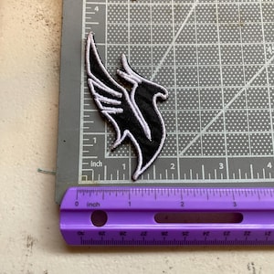 May include: An embroidered patch featuring a stylized black bird wing design with white stitching outlines. The patch is positioned on a gray cutting mat next to a purple ruler, showing the design measures approximately 2.5 inches in height.