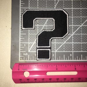 May include: A black question mark patch with a white embroidered border. The patch is positioned on a gray cutting mat with a white grid. A pink ruler is placed along the bottom edge, showing the patch is approximately 2.5 inches tall.