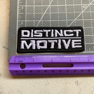 May include: A rectangular black embroidered patch featuring the white text "DISTINCT MOTIVE" in two lines separated by a horizontal line. The patch is positioned above a purple ruler, showing a width of approximately 4 inches.