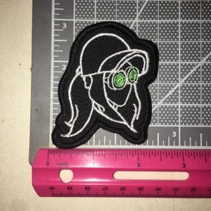 May include: A black embroidered patch featuring a white outline of a character wearing a cap and goggles with bright green lenses. The patch is positioned against a gray cutting mat with a pink ruler showing a width of approximately 3 inches.