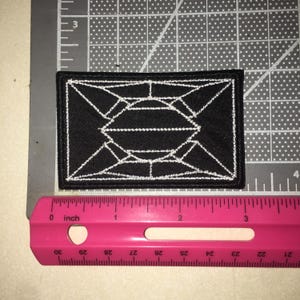 May include: A rectangular black embroidered patch featuring a white geometric line design resembling a faceted gemstone or crystal. The patch is positioned above a pink ruler, showing a width of approximately 3.5 inches and a height of 2 inches.