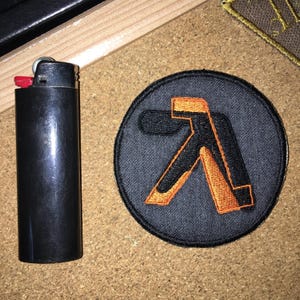 May include: A circular embroidered patch featuring a black and orange Greek lambda symbol on a dark gray background, positioned next to a standard black rectangular lighter for scale. The patch has a stitched border and a textured fabric surface.
