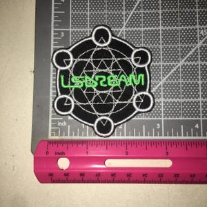May include: A black embroidered patch featuring a white geometric star pattern and the green text LSDREAM. The patch is circular with six small circles along the border and measures approximately 3 inches in width on a cutting mat.
