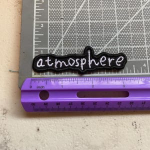 May include: A black embroidered patch with the word atmosphere in white lowercase letters. The patch is positioned on a gray cutting mat next to a purple ruler, showing the patch measures approximately 3.5 inches in total length.