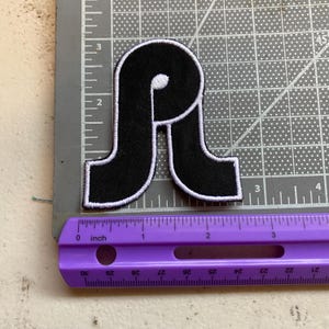 May include: A black embroidered patch in the shape of a stylized letter P with a thick white border. The patch is positioned on a gray cutting mat next to a purple ruler, showing the patch measures approximately 2.5 inches in height.