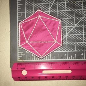 May include: A hot pink, twenty-sided die patch with white embroidered outlines. The patch is positioned on a gray cutting mat next to a pink ruler, showing the object measures approximately 2.5 inches in width and height.