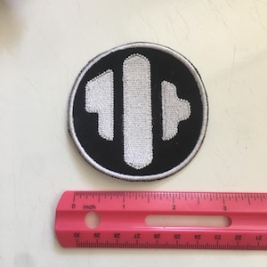 May include: A circular embroidered patch featuring a black background with a white border and a central white abstract geometric design. A pink ruler is positioned below the patch, indicating a diameter of approximately 2.5 inches.