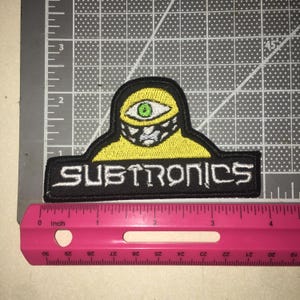May include: An embroidered patch featuring a yellow cyclops figure with a green eye and a black and white patterned mask. The word SUBTRONICS is stitched in white capital letters on a black rectangular base. A pink ruler shows it is 4 inches wide.