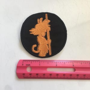 May include: A circular black embroidered patch featuring an orange silhouette of a character holding a staff. A pink ruler is positioned below the patch, showing that the circular design has a diameter of approximately 3 inches.