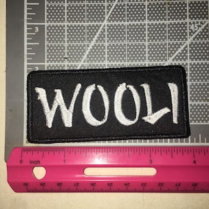 May include: A rectangular black embroidered patch featuring the white text WOOLI. The patch is positioned above a pink ruler, showing a width of approximately 4 inches and a height of approximately 1.5 inches on the grid mat background.
