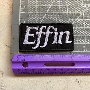 May include: A rectangular black embroidered patch featuring the white text Effin in a cursive font. The patch is positioned against a gray cutting mat with a purple ruler showing a width of approximately 3.5 inches for the embroidered design.