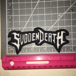 May include: A black and white embroidered patch featuring the text SUDDENDEATH in a stylized font. The patch is positioned on a cutting mat next to a pink ruler, showing the design measures approximately 5.5 inches in total width.