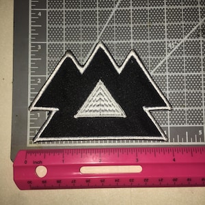 May include: A black embroidered patch in the shape of three mountain peaks with a smaller white triangle in the center. The patch is positioned against a cutting mat and a pink ruler, showing a width of approximately 4.5 inches.