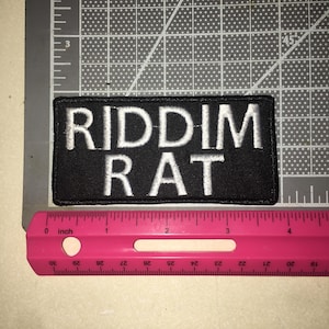 May include: A rectangular black embroidered patch with the white text RIDDIM RAT in capital letters. The patch is positioned above a pink ruler, showing a width of approximately 4 inches and a height of approximately 1.5 inches.