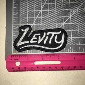 May include: A black embroidered patch featuring the word LEVITY in white script lettering. The patch is positioned on a gray cutting mat next to a pink ruler, showing the patch measures approximately 4 inches in total length from end to end.