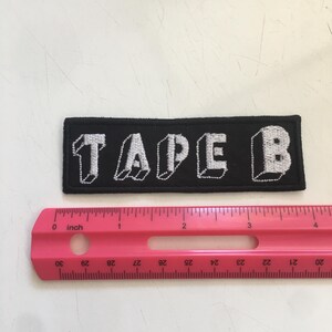 May include: A rectangular black embroidered patch featuring the white text TAPE B in a blocky, three-dimensional style. The patch is positioned above a pink plastic ruler, showing the patch measures approximately 4 inches in total length.