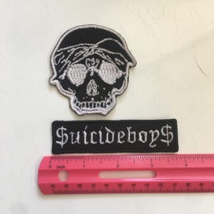 May include: Two embroidered patches with white thread on black fabric. One patch is a skull wearing a bandana and sunglasses. The other is a rectangular patch with the text $uicideboy$ in a gothic font. A pink ruler shows the scale in inches.