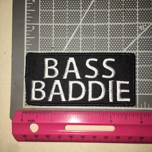 May include: A rectangular black embroidered patch with white text reading BASS BADDIE. The patch is positioned on a gray cutting mat next to a pink ruler, showing the patch measures approximately 4 inches in length and 1.5 inches in height.