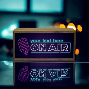May include: A wooden framed sign with a neon pink microphone and blue neon text that reads "your text here ON AIR" on a brick background.
