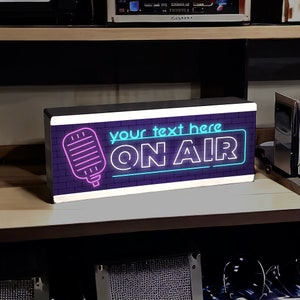 May include: A black and white neon sign with a pink microphone and blue text that says "your text here ON AIR".
