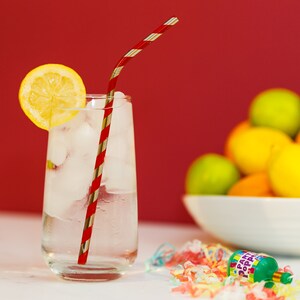 May include: A glass of ice water with a red and silver striped straw. The glass is on a white surface with a lemon wedge and a party popper with confetti.