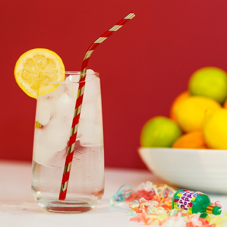 May include: A glass of ice water with a red and silver striped straw. The glass is on a white surface with a lemon wedge and a party popper with confetti.