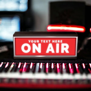 May include: A red and black sign with the words "YOUR TEXT HERE ON AIR" in white letters. The sign is sitting on a black surface in front of a keyboard.