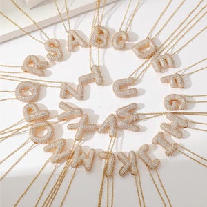 May include: A set of gold necklaces with letter pendants. Each pendant is made of clear cubic zirconia stones and is shaped like a letter of the alphabet. The necklaces are all different lengths and are arranged in a circle on a white background.