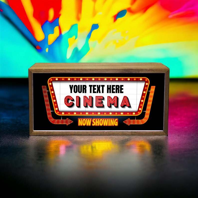 Puede incluir: Un letrero de madera con un marquesina roja y blanca que dice "YOUR TEXT HERE CINEMA" y "NOW SHOWING". El letrero est&aacute; sobre una superficie oscura con un fondo colorido.