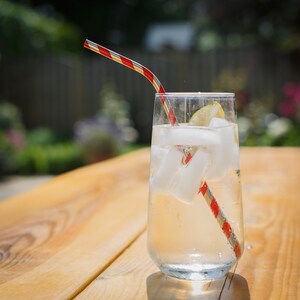 May include: A glass of ice water with a lime wedge and a red and white striped straw on a wooden table.