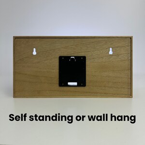 Pu&ograve; includere: Il retro di un cartello in legno con due buchi per l'appeso e un vano batteria nero. Il cartello dice "Self standing or wall hang".