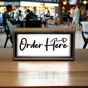May include: A wooden sign with a white background and black text that reads "Order Here" with an arrow pointing to the right. The sign is sitting on a wooden table.