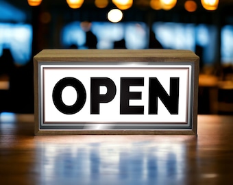 May include: A wooden sign with a white background and black lettering that reads "OPEN". The sign is illuminated and sits on a wooden table.