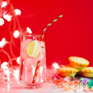 May include: A tall glass filled with ice and a lime wedge, with a red and white striped straw. The glass is on a white surface with a red background and festive confetti.